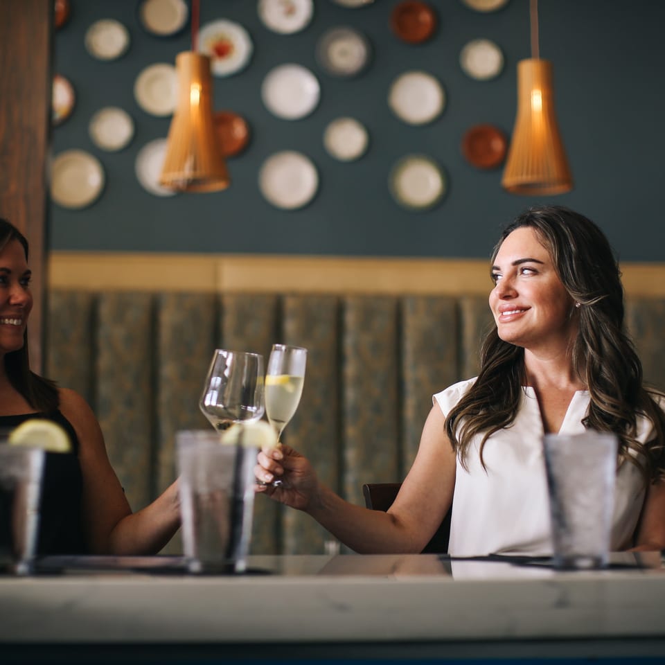 Two women toast with wine at a warmly lit restaurant table, surrounded by pendant lights and decorative plates.