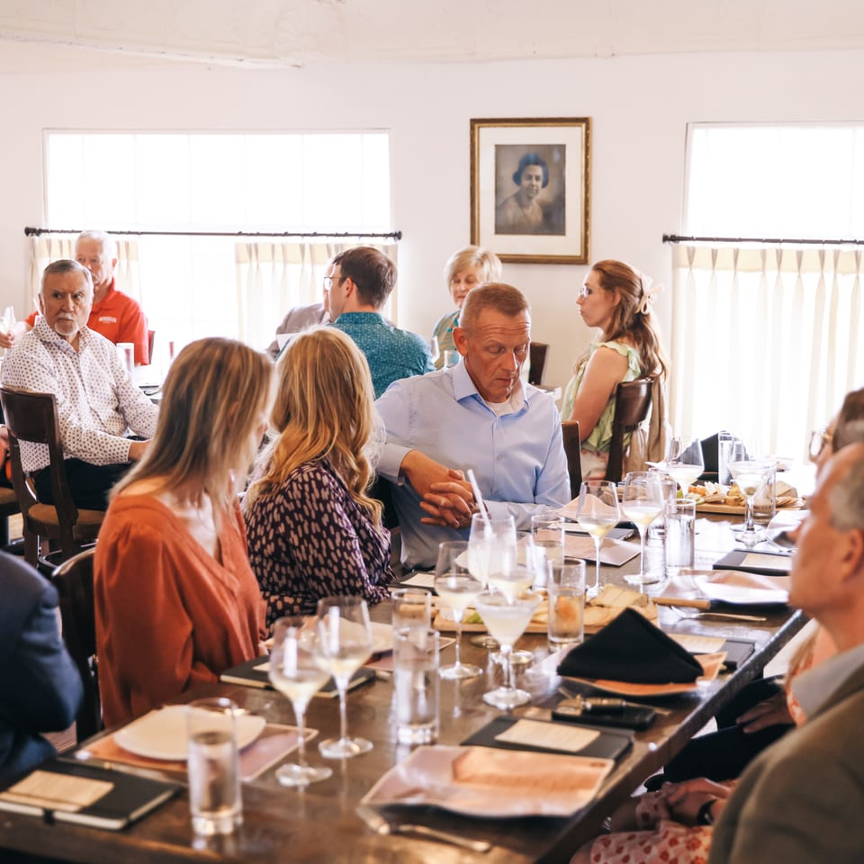 Large group dining together at a warmly lit restaurant, with wine glasses, plated meals, and a standing server.