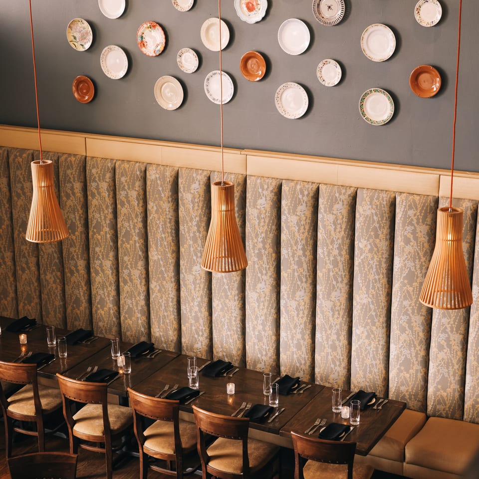Elegant restaurant dining room with patterned banquette seating, pendant lights, set tables, and decorative plates displayed on the wall.