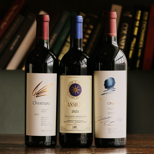 Three bottles of red wine, Overture, Sassicaia, and Opus One, stand on a wooden table.