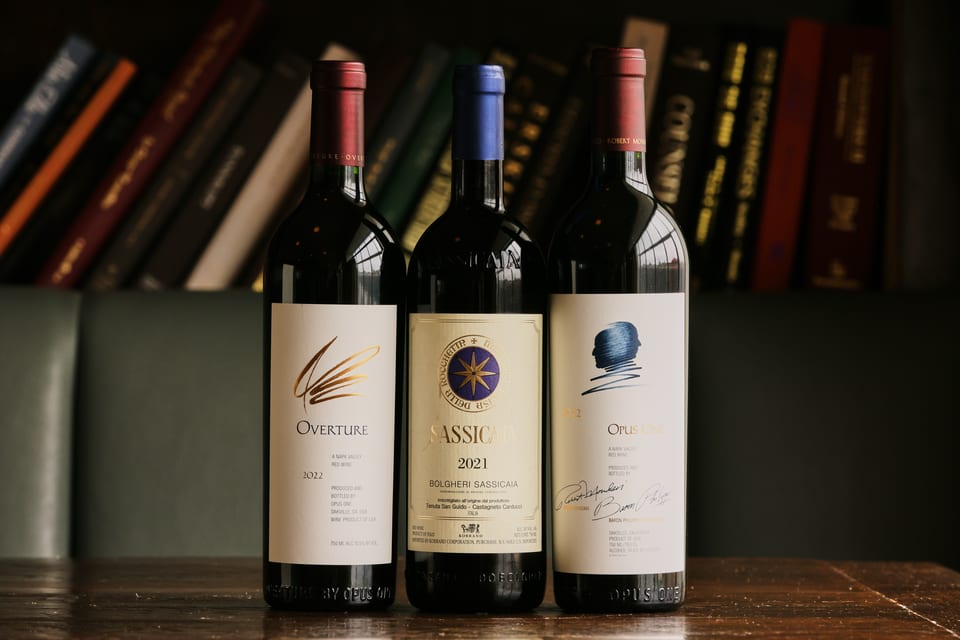 Three bottles of red wine, Overture, Sassicaia, and Opus One, stand on a wooden table.