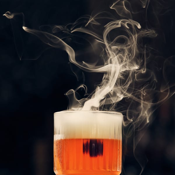A vibrant orange cocktail with a white foam top, emitting dramatic smoke against a dark background.