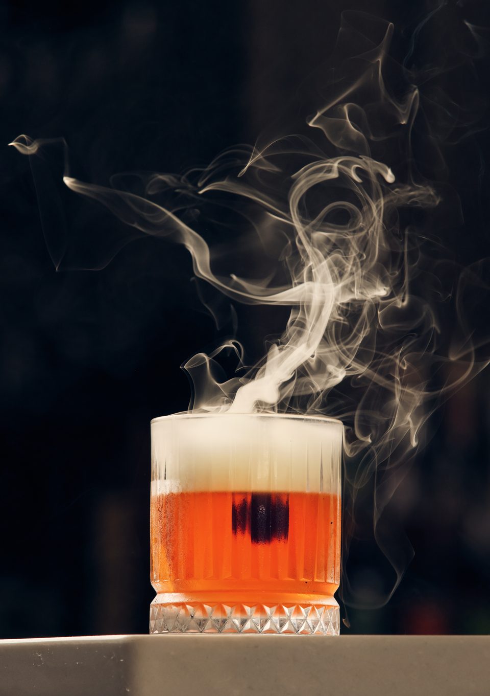 A vibrant orange cocktail with a white foam top, emitting dramatic smoke against a dark background.