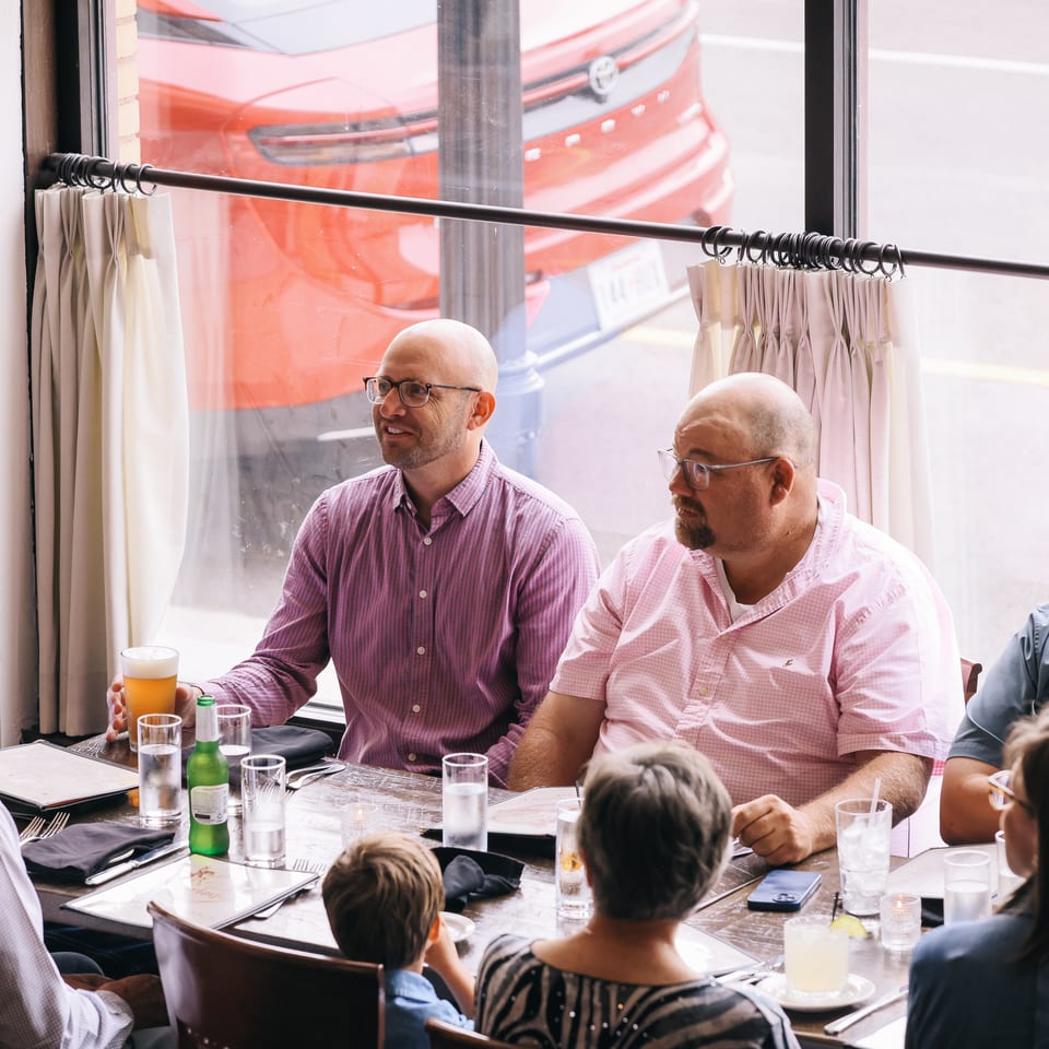 Group of diners enjoying drinks and conversation around a restaurant table beside a large street-facing window.