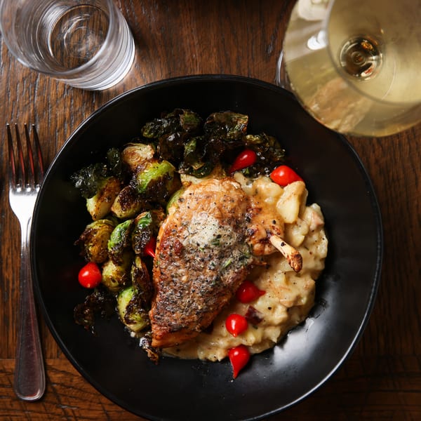 Crispy chicken with roasted Brussels sprouts, mashed potatoes, and cherry tomatoes on a dark plate.