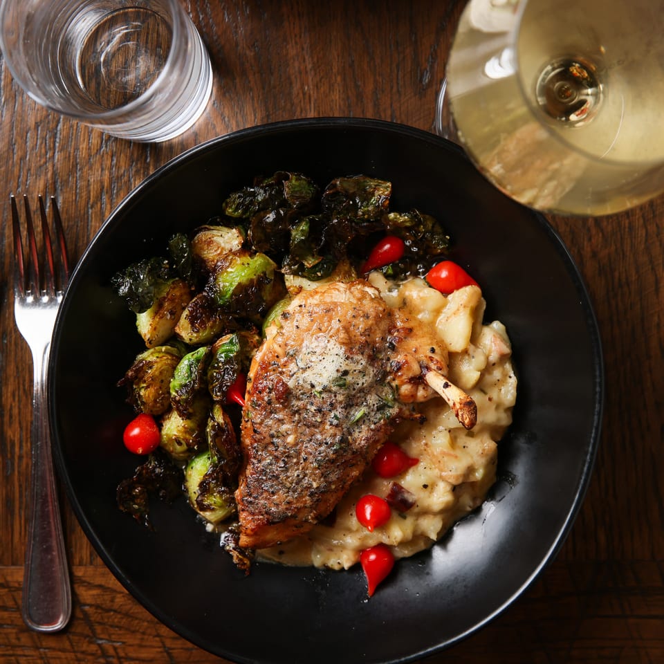 Crispy chicken with roasted Brussels sprouts, mashed potatoes, and cherry tomatoes on a dark plate.
