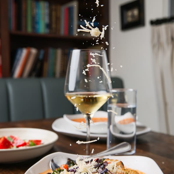 A hand sprinkles shredded cheese over a bowl of shrimp pasta with a glass of white wine.