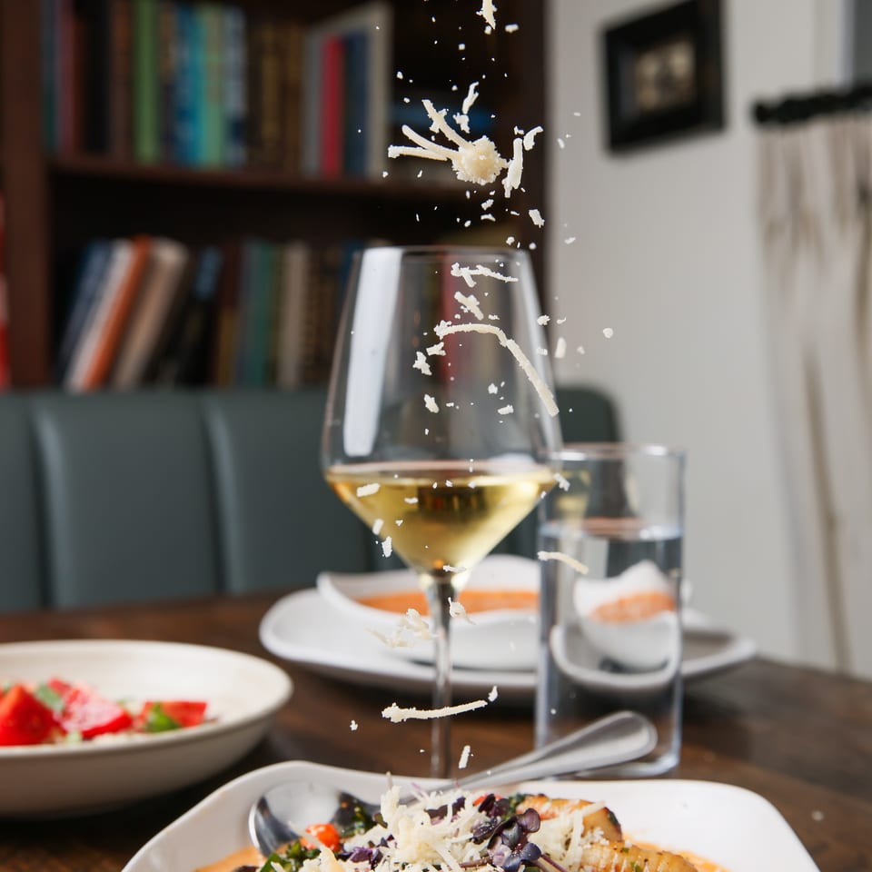 A hand sprinkles shredded cheese over a bowl of shrimp pasta with a glass of white wine.