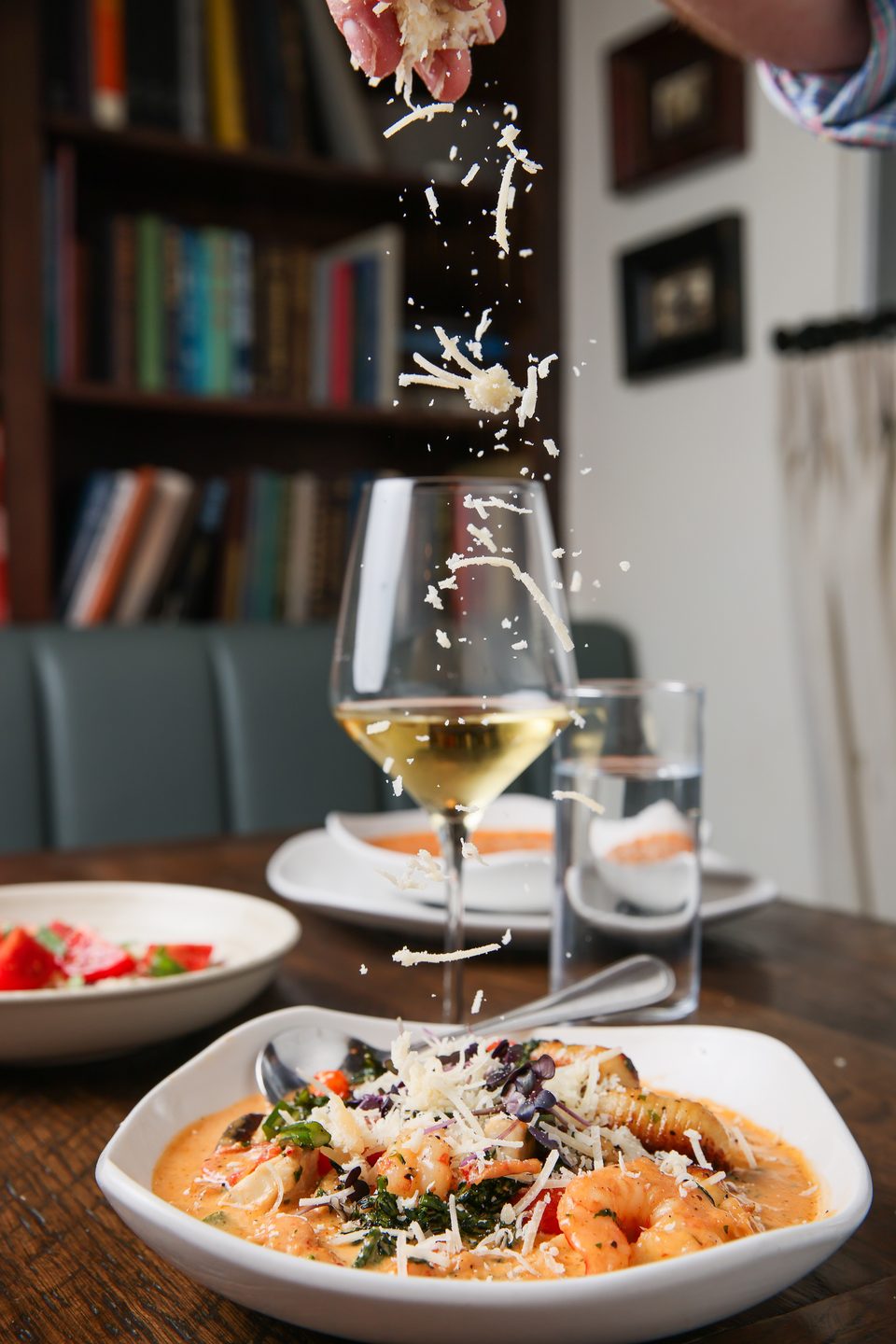 A hand sprinkles shredded cheese over a bowl of shrimp pasta with a glass of white wine.