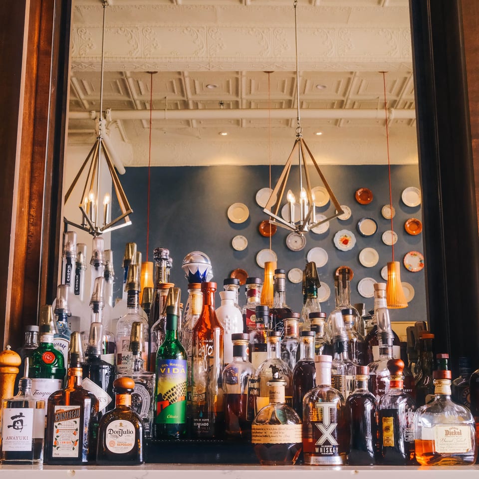 Restaurant bar stocked with liquor bottles, mirrored walls, decorative plates, and geometric pendant lights.