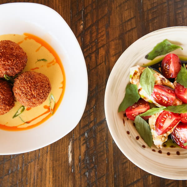 Two dishes on a wooden table: fried arancini in sauce and a caprese salad with tomatoes, mozzarella, and basil.