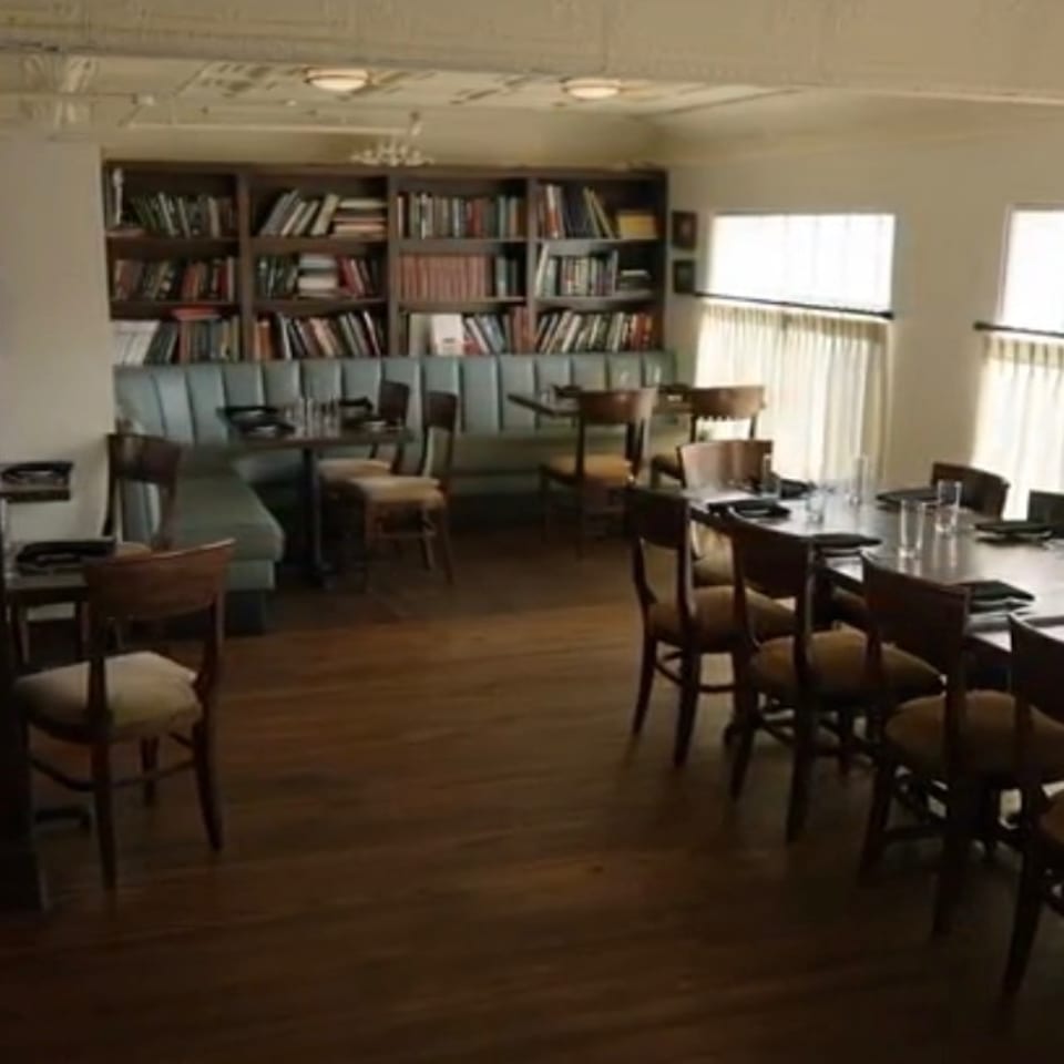Sunlit restaurant dining room with wooden tables, upholstered chairs, banquette seating, and bookshelves lining the back wall.